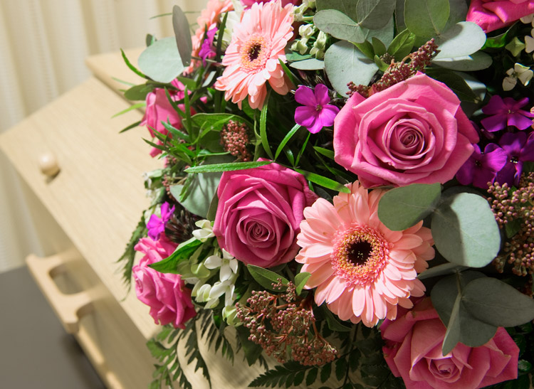 Bouquet of pink roses and daisies with green leaves rests atop a light-colored wooden casket, situated against a background of cream-colored curtains.